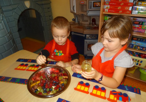 Dwoje dzieci siedzi przy stole. Dziewczynka trzyma w ręku słoik z miodem i obserwuje chłopca który łyżką miesza miód z pokrojonymi owocami.